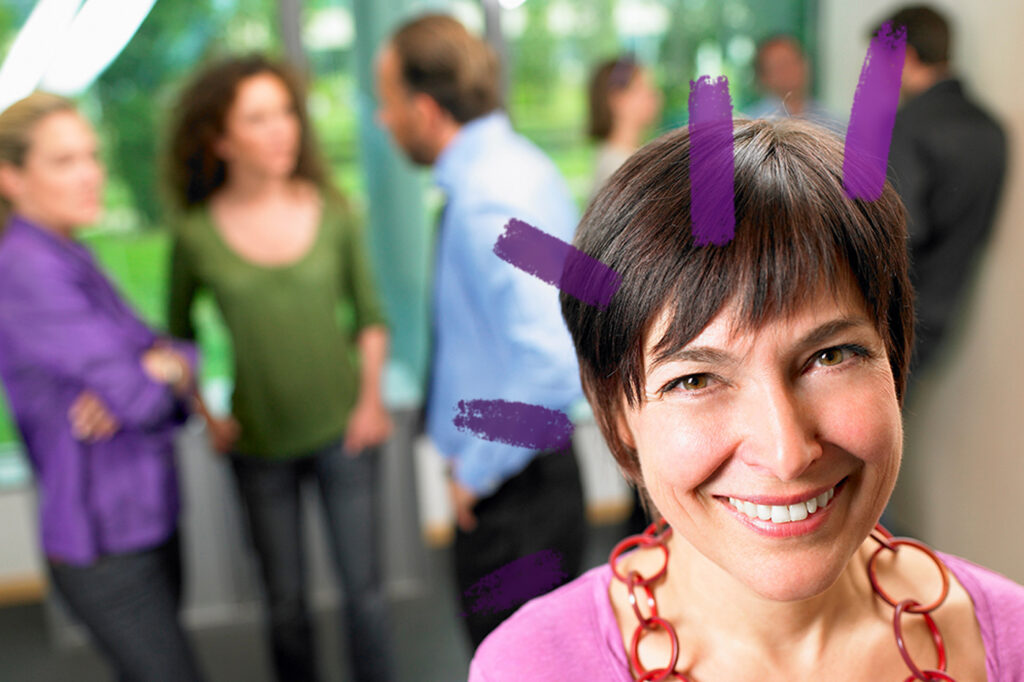 Woman at a professional event smiling at the camera. In the background work colleagues are mingling.